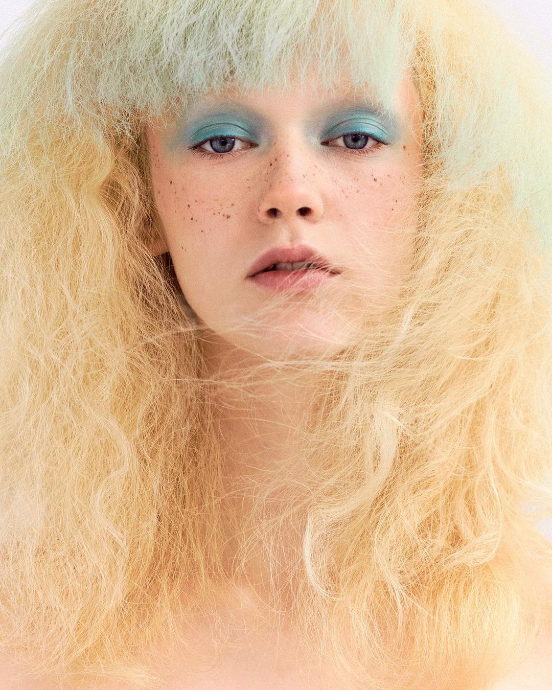 Close-up portrait of a female with blonde, textured hair, blue eyeshadow, and freckles against a white background, beauty shot.