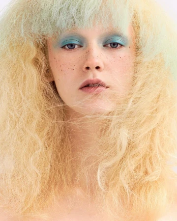 Close-up portrait of a female with blonde, textured hair, blue eyeshadow, and freckles against a white background, beauty shot.