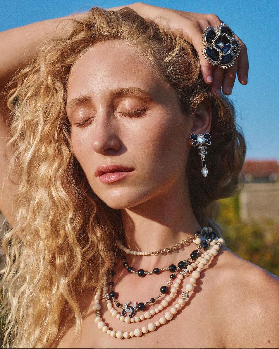 Woman with blonde hair, wearing jewelry, including a ring and layered necklaces. She poses against a blue sky background.