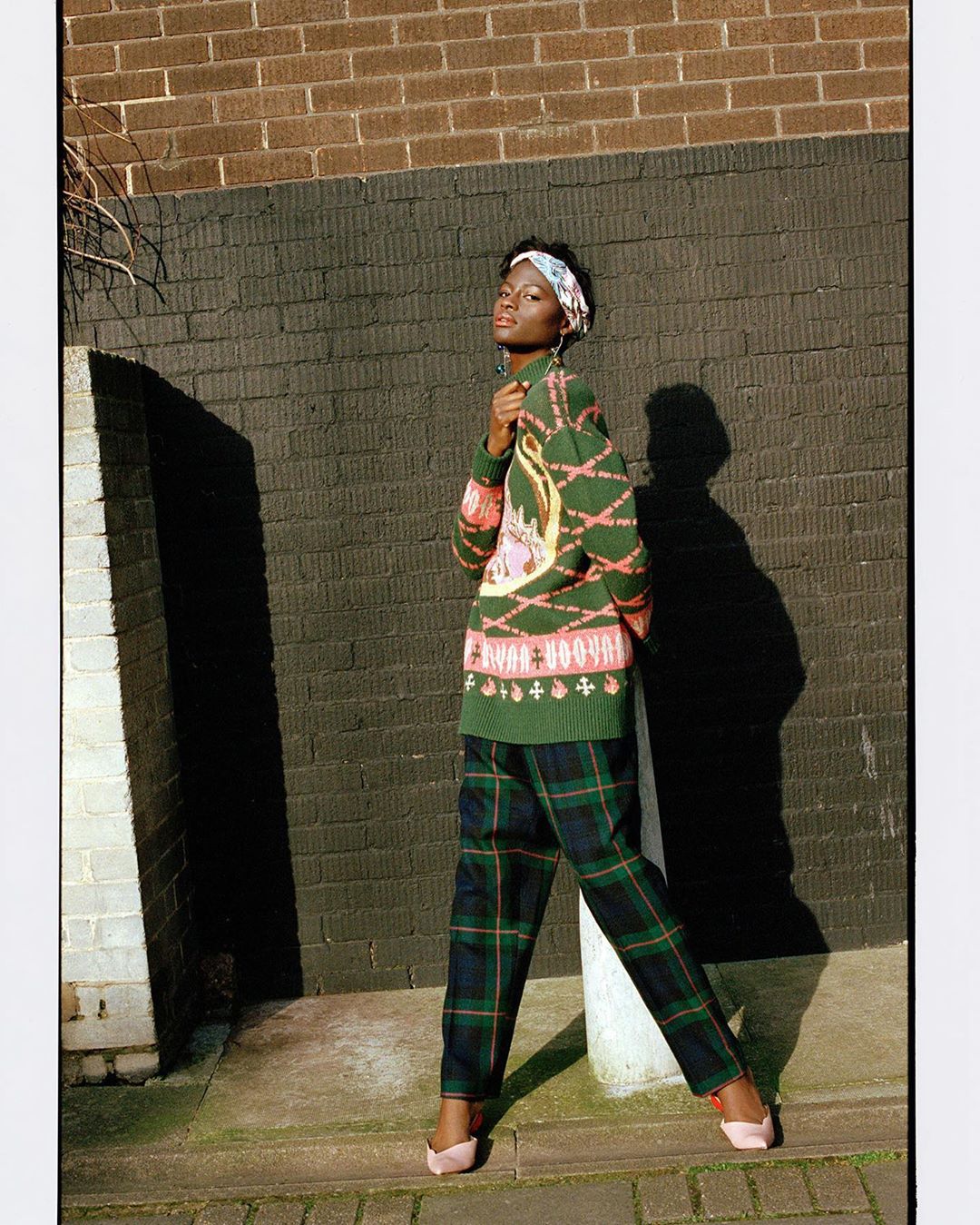 A woman posing outdoors in a patterned green sweater and plaid pants, standing against a brick wall on a sunny day for fashion editorial.
