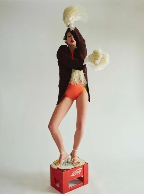 Fashion editorial photo of a woman in an orange bodysuit and blazer, standing on a red crate, holding feather dusters against a white backdrop.