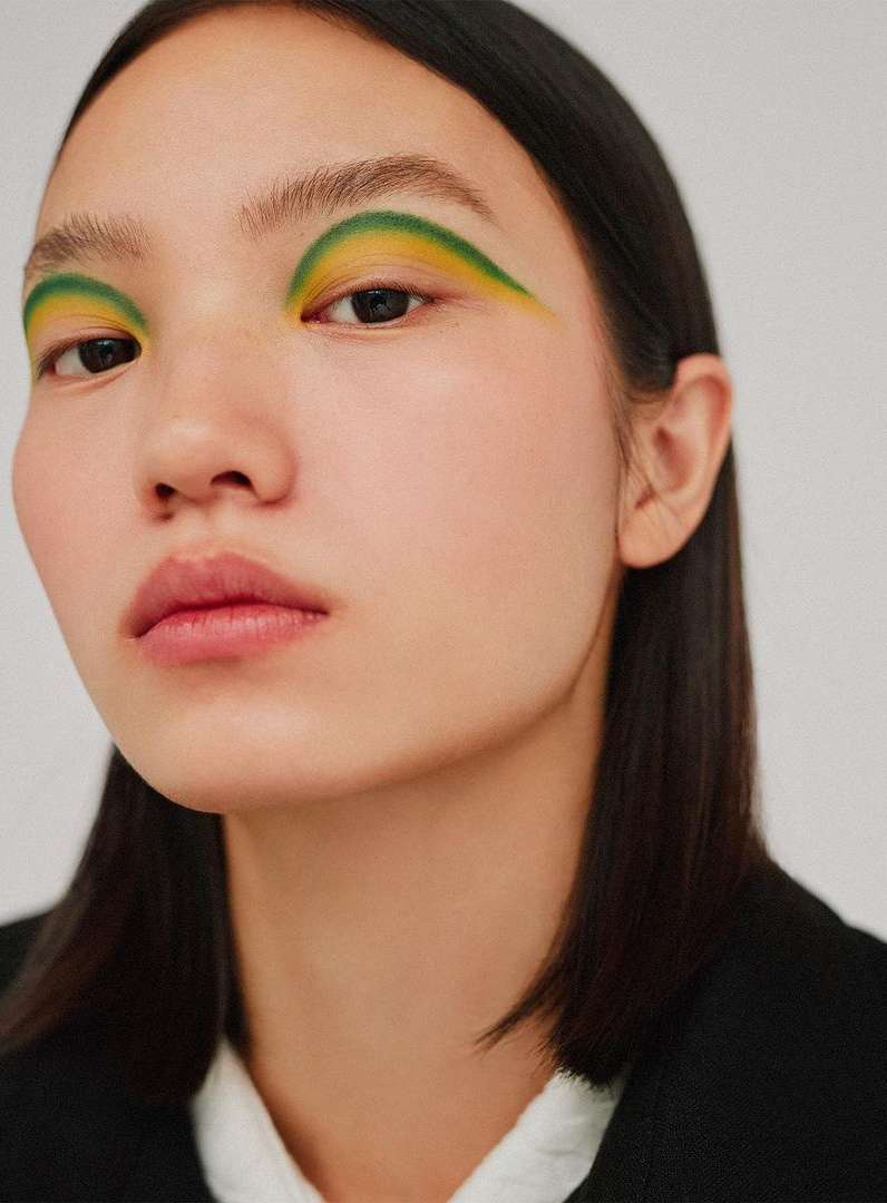 Close-up studio portrait of an Asian adult female with vibrant green and yellow eye makeup in a beauty and fashion context.