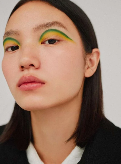 Close-up studio portrait of an Asian adult female with vibrant green and yellow eye makeup in a beauty and fashion context.