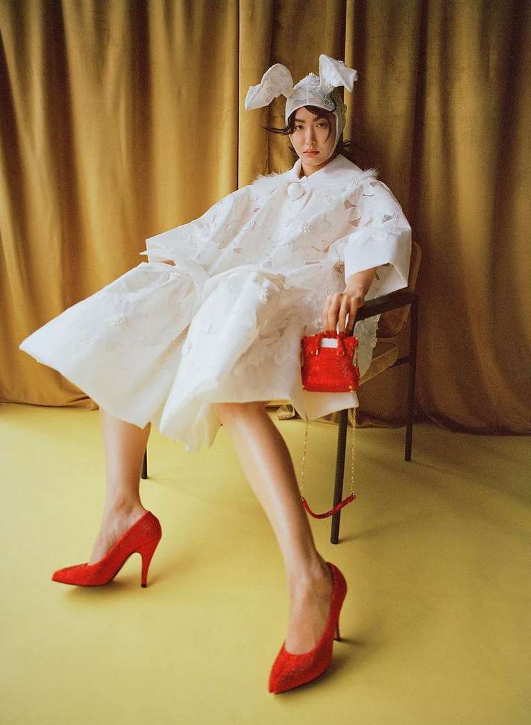 A woman in a white dress, bunny hat, and red heels sits on a chair holding a small red purse against a yellow backdrop.