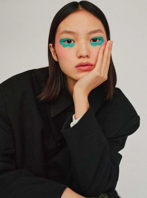 Close-up of a young woman with green cloud-shaped eyeshadow and dark hair, resting her face on her hand against a plain background.