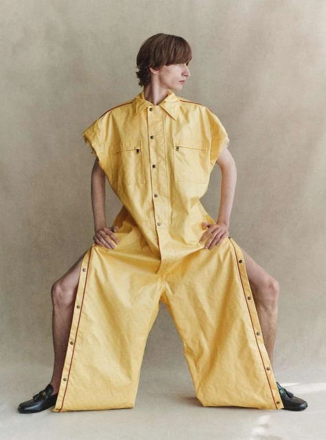 A person in a wide-leg yellow jumpsuit with short sleeves sits with legs spread, wearing black dress shoes in a neutral studio setting.
