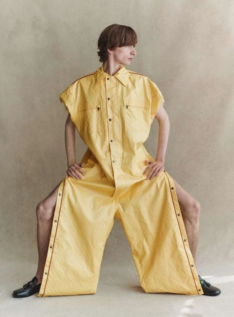 Editorial fashion shot of a young adult with long brown hair in an oversized yellow utility suit posing in front of a neutral background.