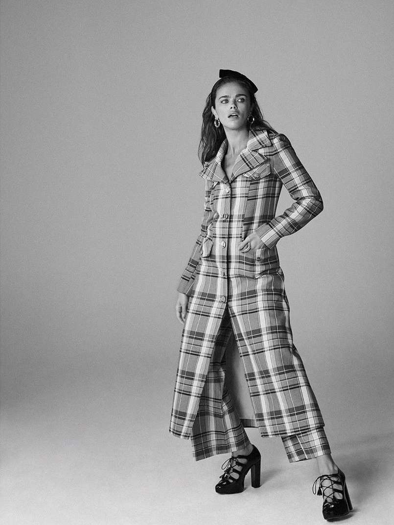 A woman in a plaid coat and platform heels poses in a studio. The black-and-white shot features a full-length view against a plain background.