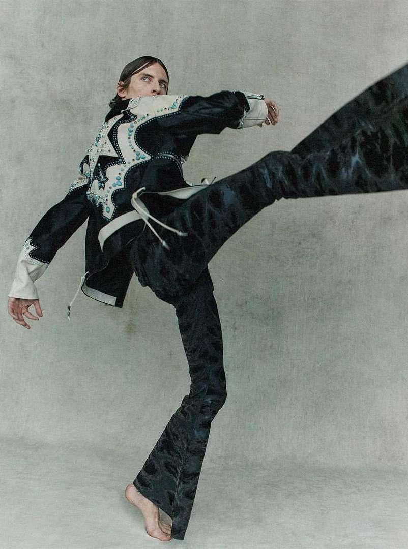 Adult male in motion wearing a decorated black jacket and flared black pants in a light grey photo studio setting, fashion editorial.