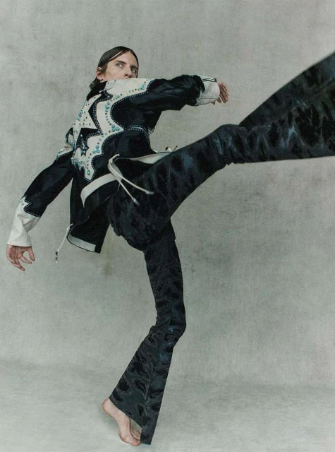 Adult male in motion wearing a decorated black jacket and flared black pants in a light grey photo studio setting, fashion editorial.