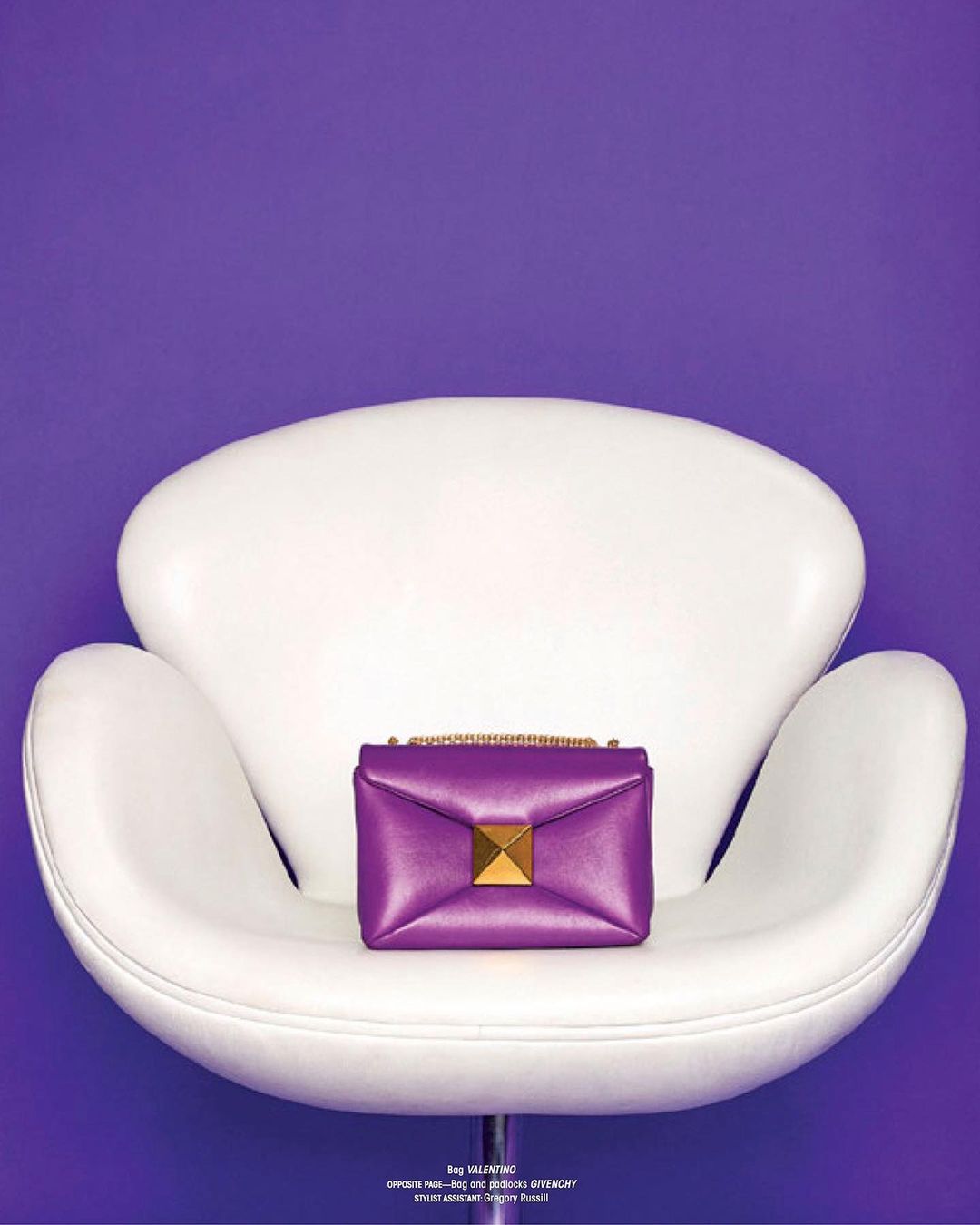 Purple leather Valentino handbag with a gold pyramid stud closure sits on a vintage white chair against a solid purple background.