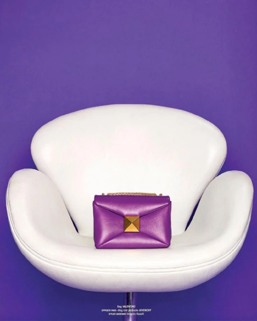 Purple leather Valentino handbag with a gold pyramid stud closure sits on a vintage white chair against a solid purple background.