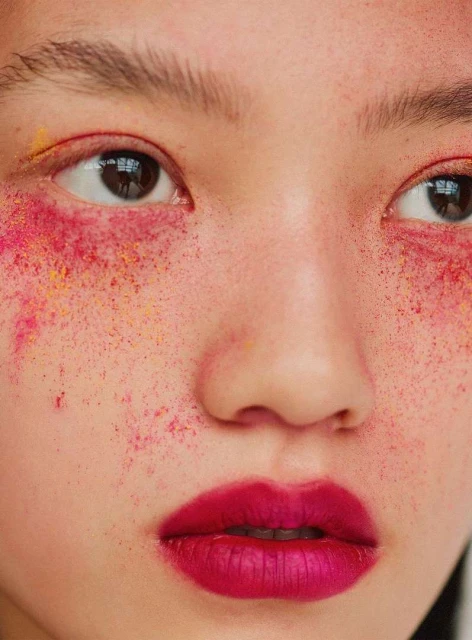 Close-up beauty shot of a young adult female with vibrant pink lipstick and colorful cosmetic face powder, set indoors with soft lighting.