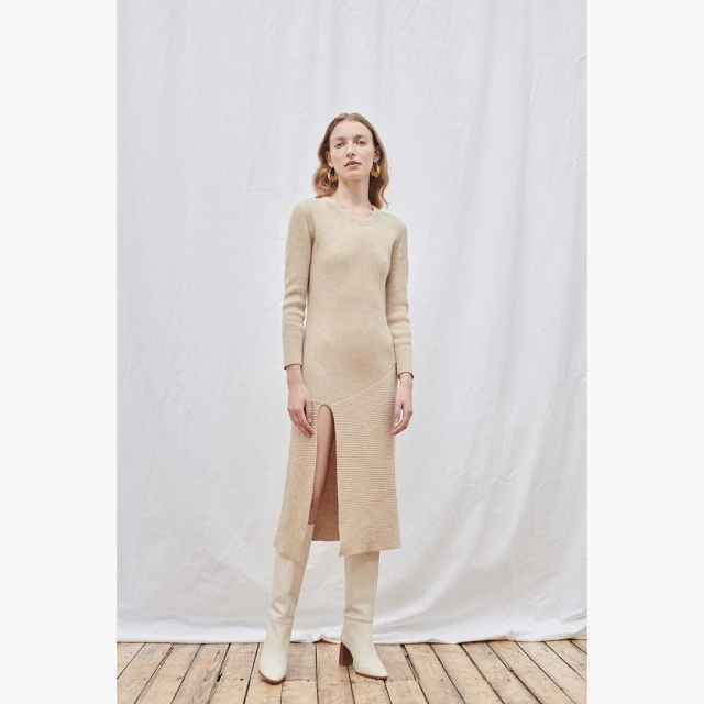 A woman models a long-sleeved cream-colored midi dress with a thigh-high slit and matching heeled boots against a neutral backdrop.