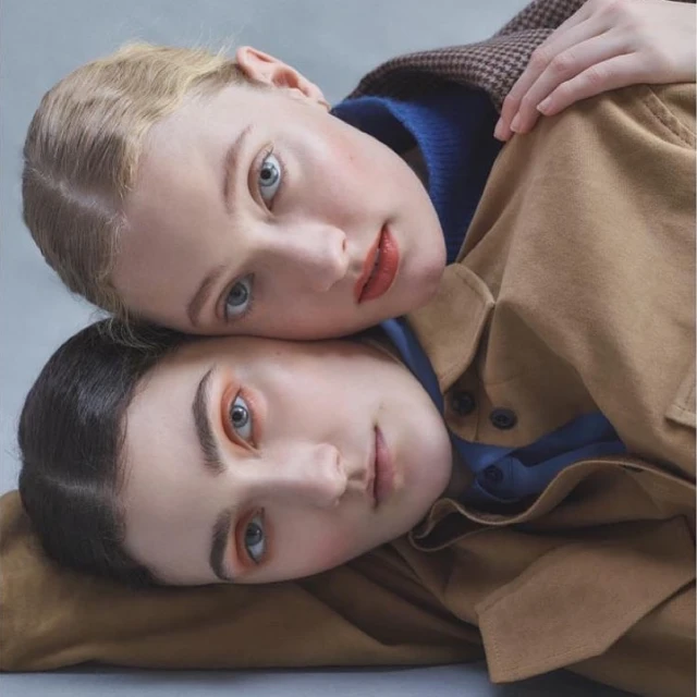 Two young women with fair skin pose together indoors wearing neutral-toned jackets, showcasing a close-up shot with soft lighting and muted colors.