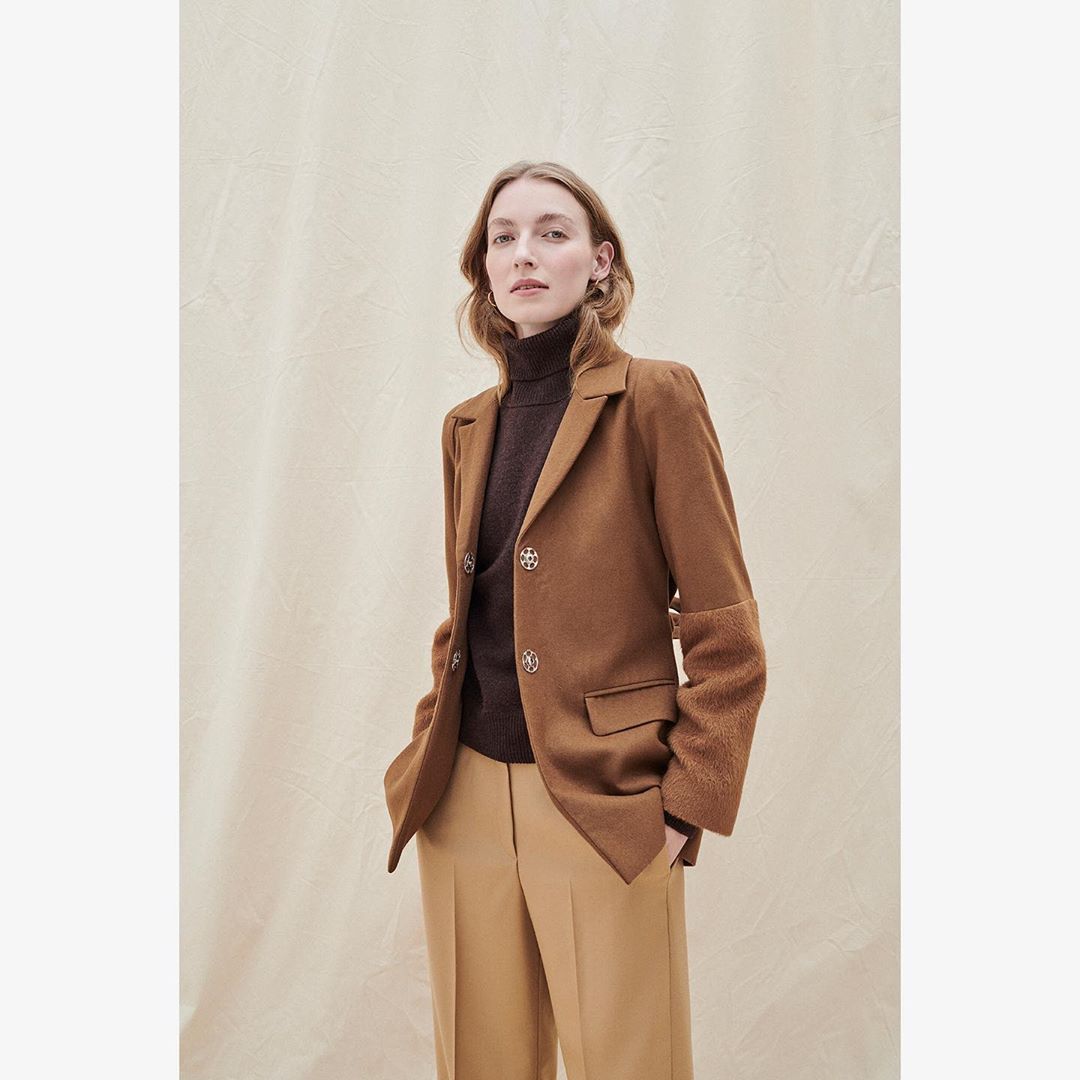Woman in brown blazer and tan trousers posing in front of a neutral background, indoor studio lighting, fashion editorial photography.