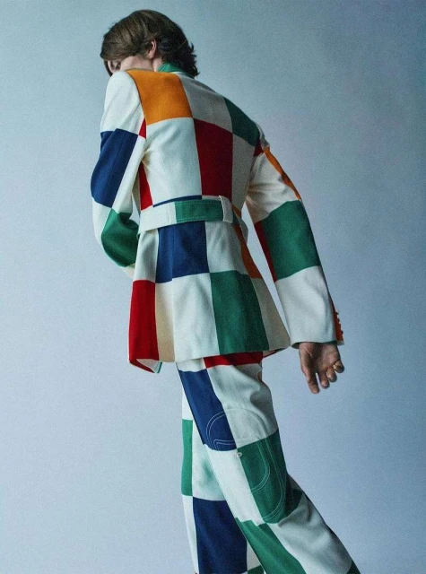 Person in a colorful patchwork suit with a belt poses in front of a light background, showcasing fashion and design.