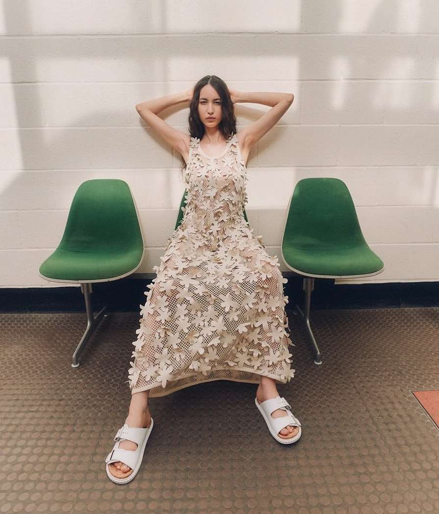 A woman in a floral dress poses seated between green chairs, in bright, natural light in an indoor environment with a light-colored background.