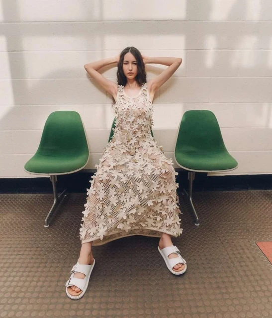 A woman in a floral dress poses seated between green chairs, in bright, natural light in an indoor environment with a light-colored background.