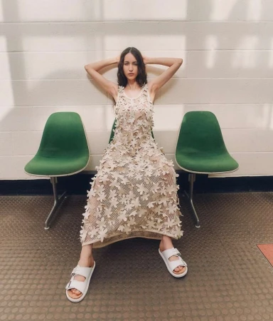 A woman in a floral dress poses seated between green chairs, in bright, natural light in an indoor environment with a light-colored background.
