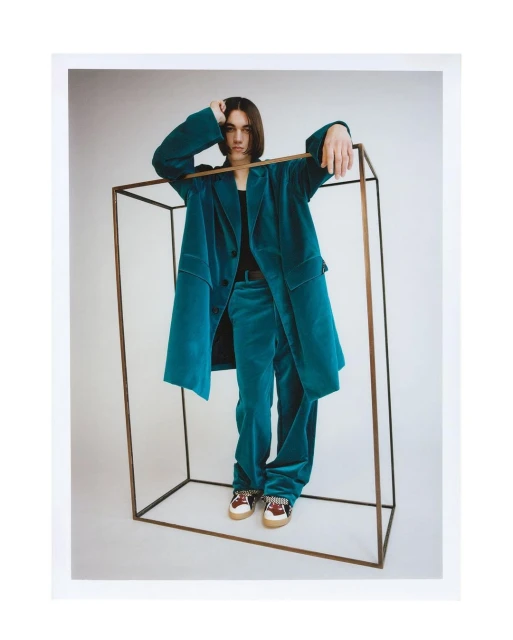 Young adult wearing a teal velvet suit, standing in the center of a dark metal cube against a plain light background in a studio.