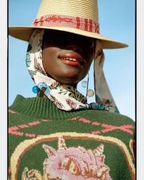 Model wearing a straw hat with patterned scarf, dangling earrings, and a green patterned sweater against a clear sky background