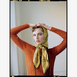 Woman wearing a yellow patterned silk headscarf and ribbed rust orange cardigan against a plain wall.