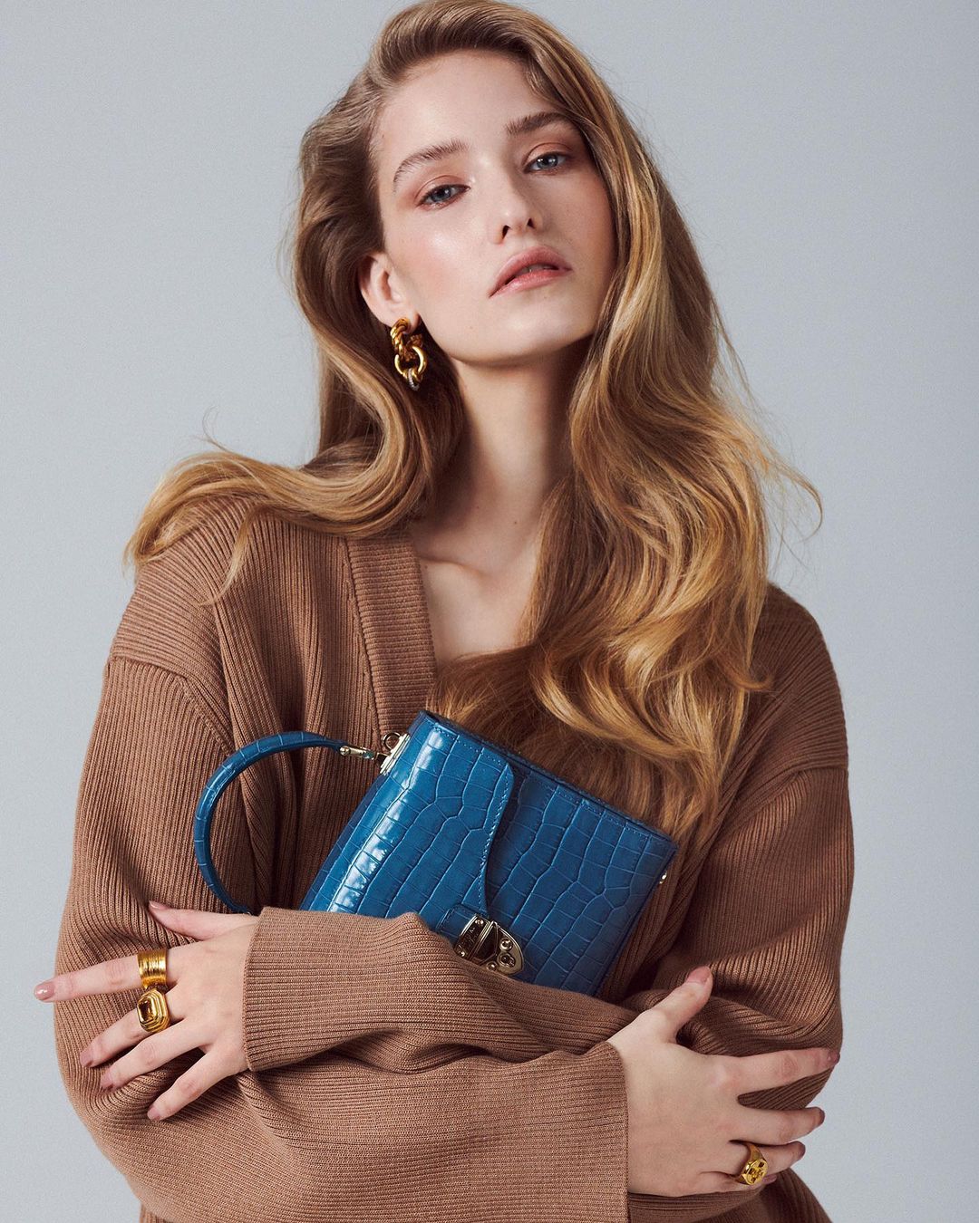 Woman in brown knit cardigan holding a blue handbag, photographed by Christopher Fenner against a plain grey background.