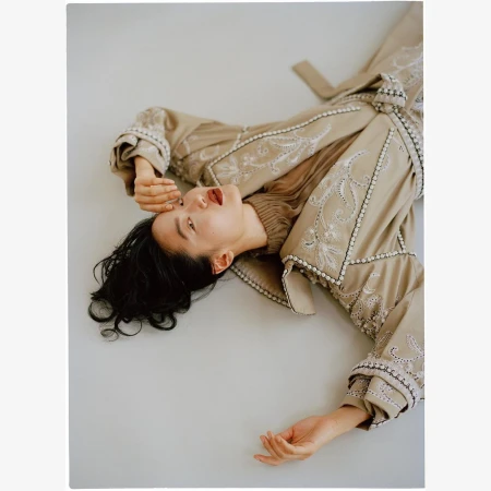 Model lying on back wearing a tan embroidered coat and dark lipstick against a neutral studio background