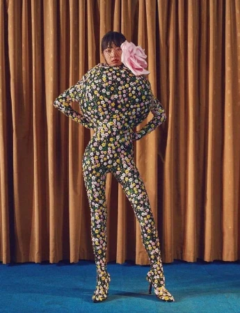 Model in a floral print catsuit with a spherical torso and pink hair flower standing against brown curtains.