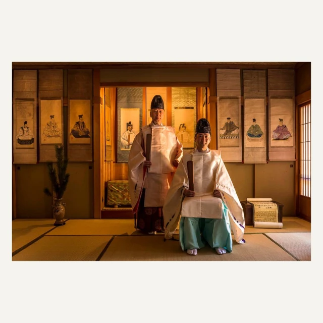 Two adult men in traditional Japanese clothing seated and standing indoors with scroll paintings, neutral colors, and soft, diffused light.