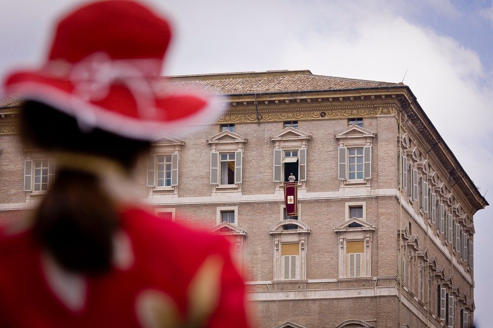 Distant view of a person in a window of a tall building, with a blurred figure in red and white in the foreground.