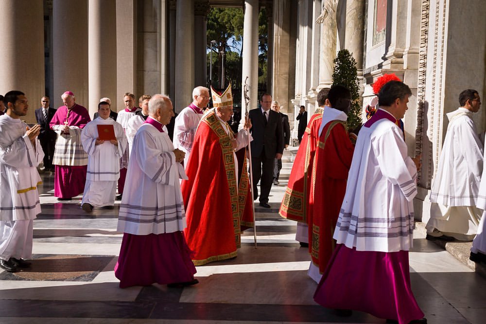 Procession of clergy walking outside a building with columns, wearing white and red robes and hats, sunny day.