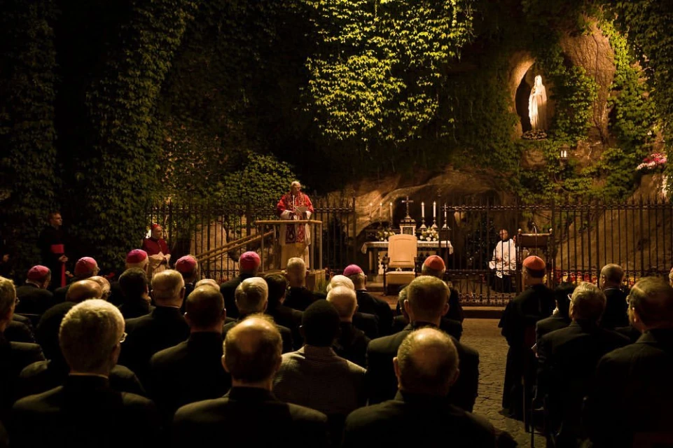 Outdoor religious gathering at night with a robed speaker, audience, statue, and greenery in dim light, creating a solemn atmosphere.