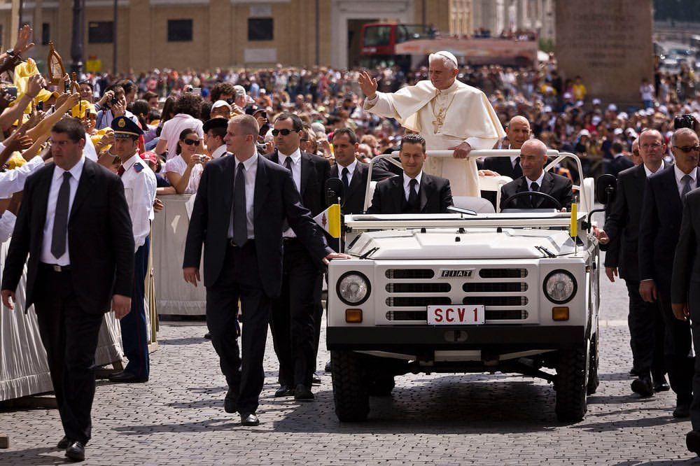 Pope waves from white Fiat with security in crowd. Daytime outdoor scene in Vatican City. People in suits flank vehicle, many bystanders.