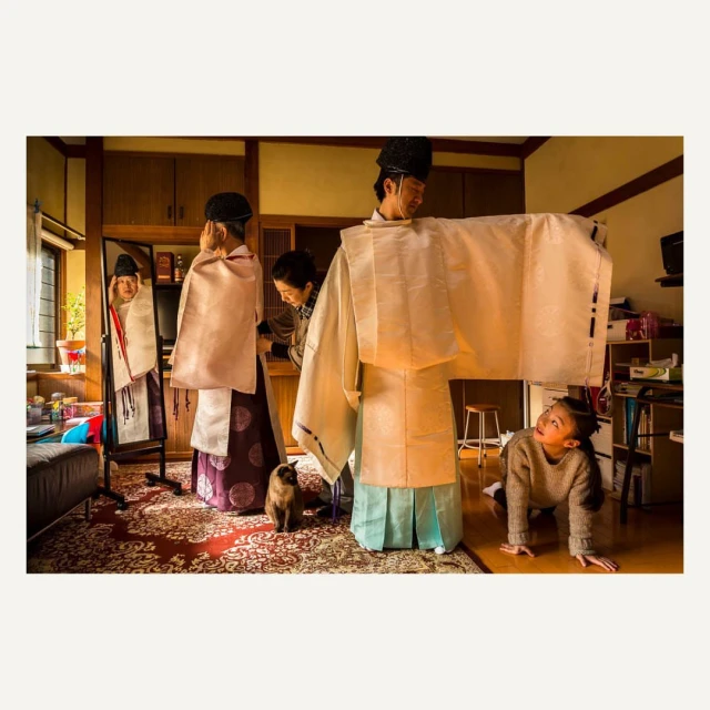 Family prepares in traditional Japanese clothing in a warm indoor setting. A young girl looks on from the floor near a Siamese cat.