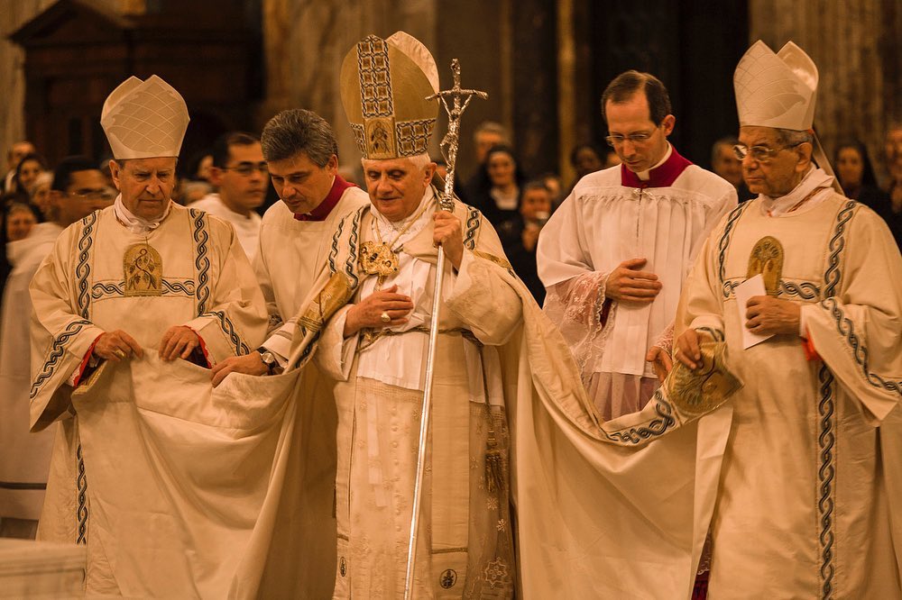 A group of men in religious attire, including a man with a cross staff, stand together indoors in a formal setting with a large group visible in the background.