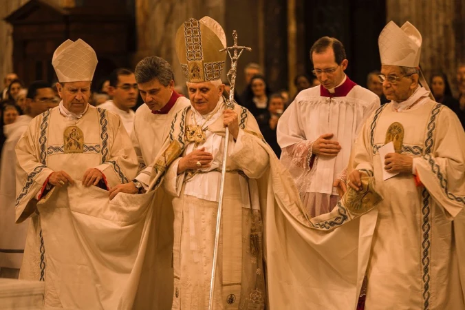 A group of men in religious attire, including a man with a cross staff, stand together indoors in a formal setting with a large group visible in the background.