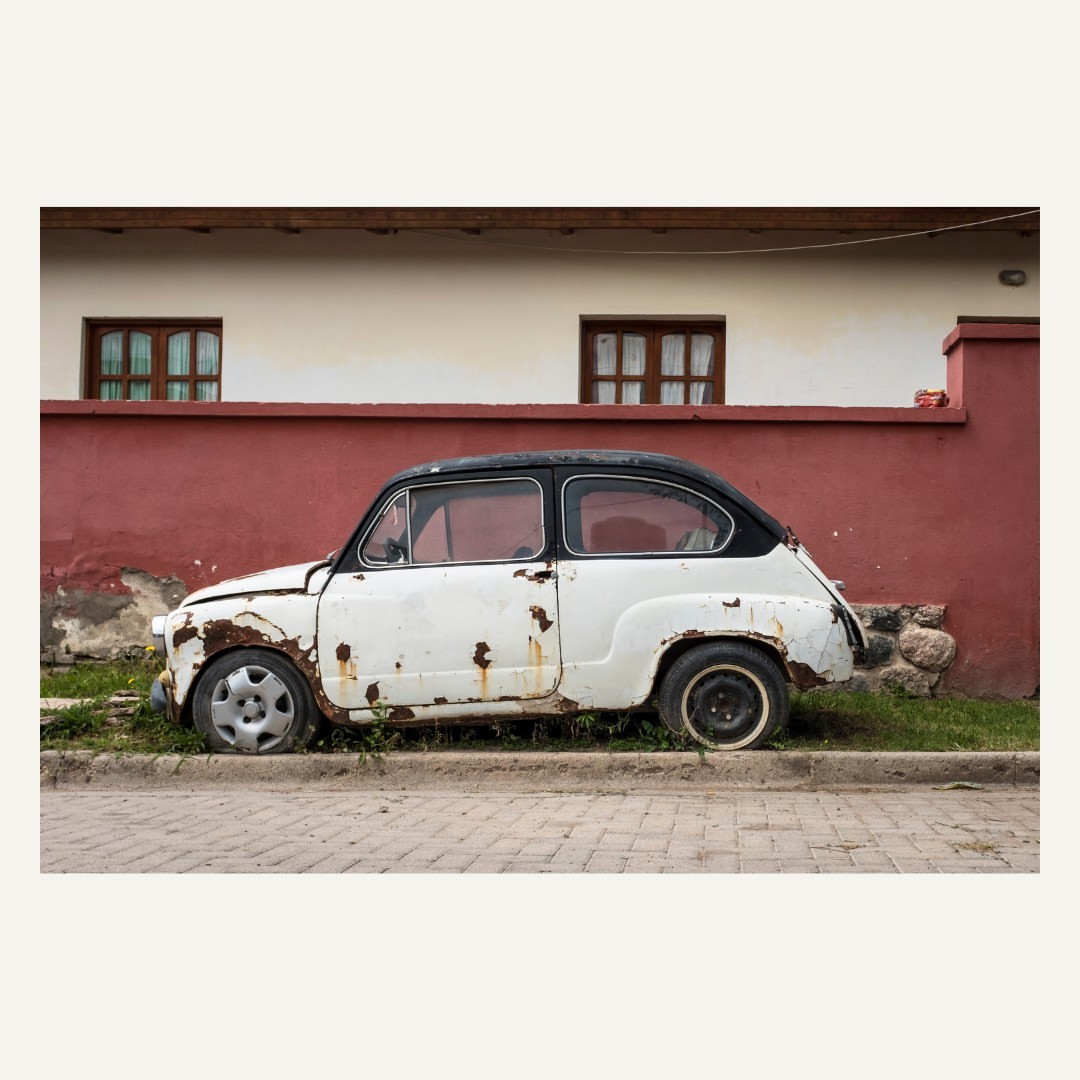 Vintage white and black car parked beside a red and white building with two windows, conveying a sense of timeless decay.