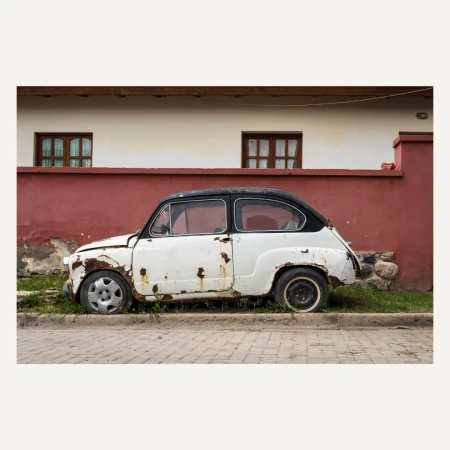 Vintage white and black car parked beside a red and white building with two windows, conveying a sense of timeless decay.