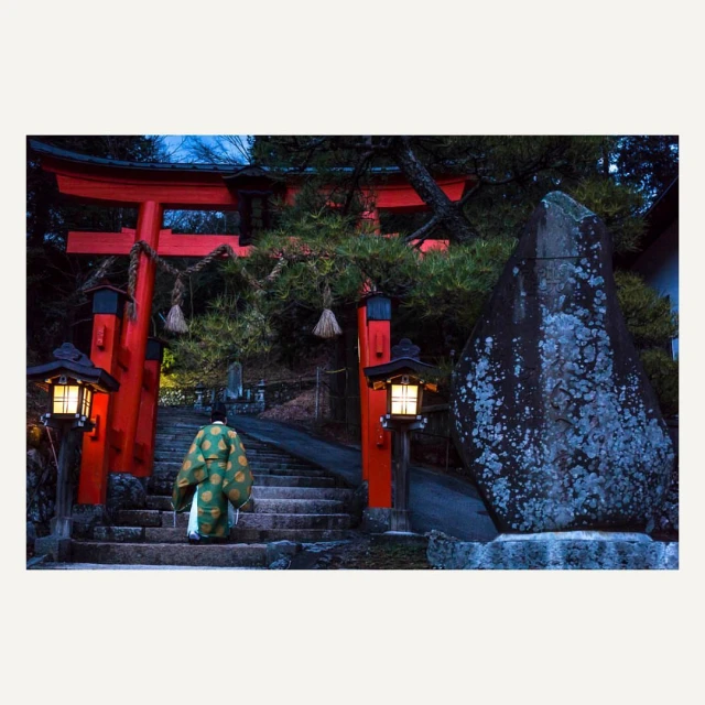 Man in traditional Japanese clothing ascends stone steps toward a red torii gate, illuminated by lanterns, in a natural setting.
