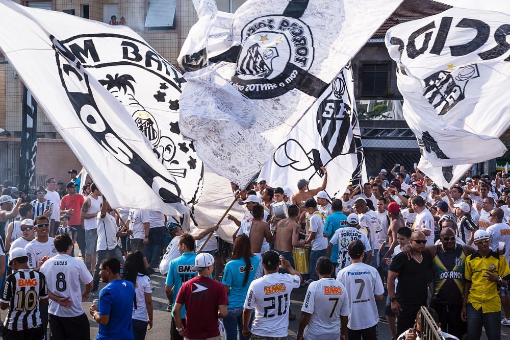 Large crowd of soccer fans wave black and white flags in the street during a daytime celebration with buildings visible in background.