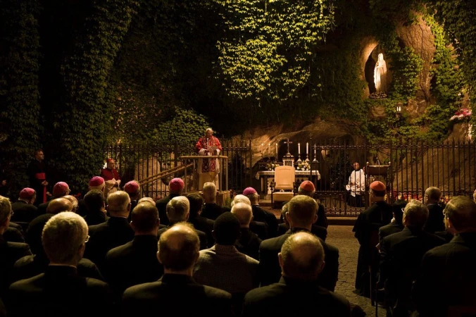 Outdoor gathering with a religious leader at a podium addressing a crowd near a grotto with a statue of the Virgin Mary, dark lighting.