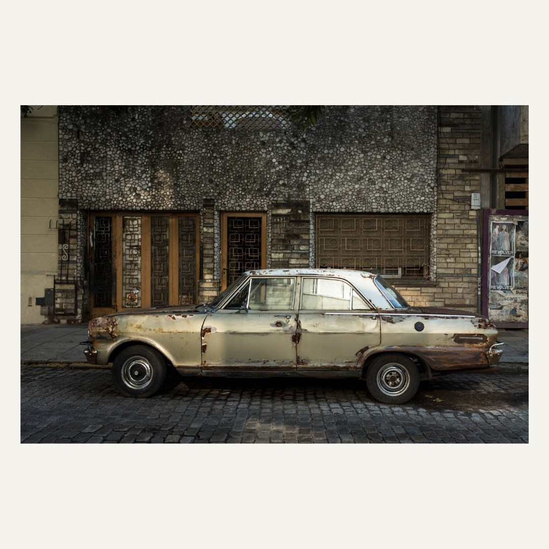 Vintage, weathered car parked on a cobblestone street in front of a textured building with wooden doors and window shutters.