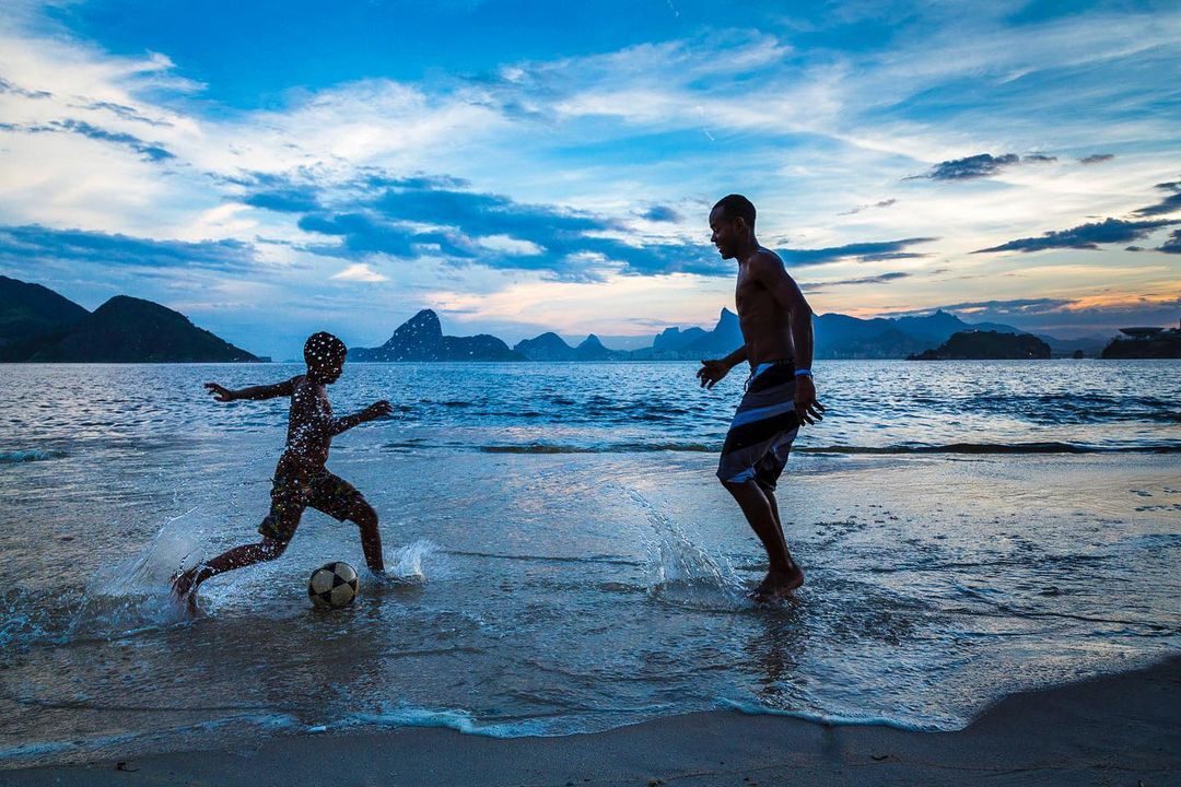 Two people play soccer on a beach with waves and mountains in the background under a cloudy blue sky at dusk or dawn.