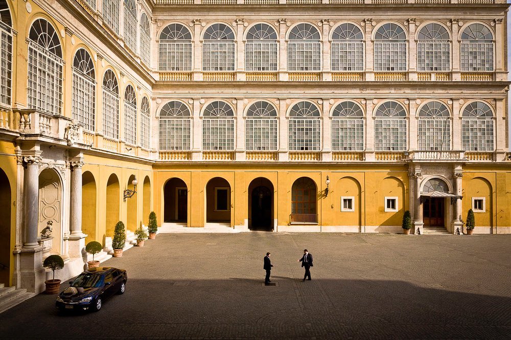 Italian architecture with two men in suits standing in a courtyard, framed by arched walkways and a black car under tall windows.