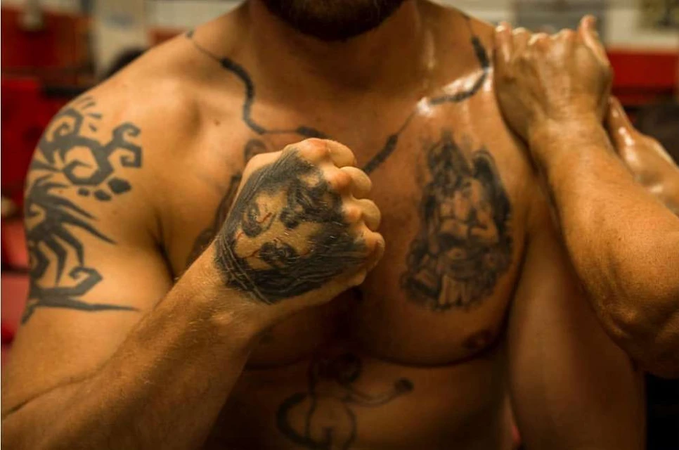 Torso of muscular adult male with tattoos on arms, chest, and hand, photographed in a fitness setting with neutral color palette.