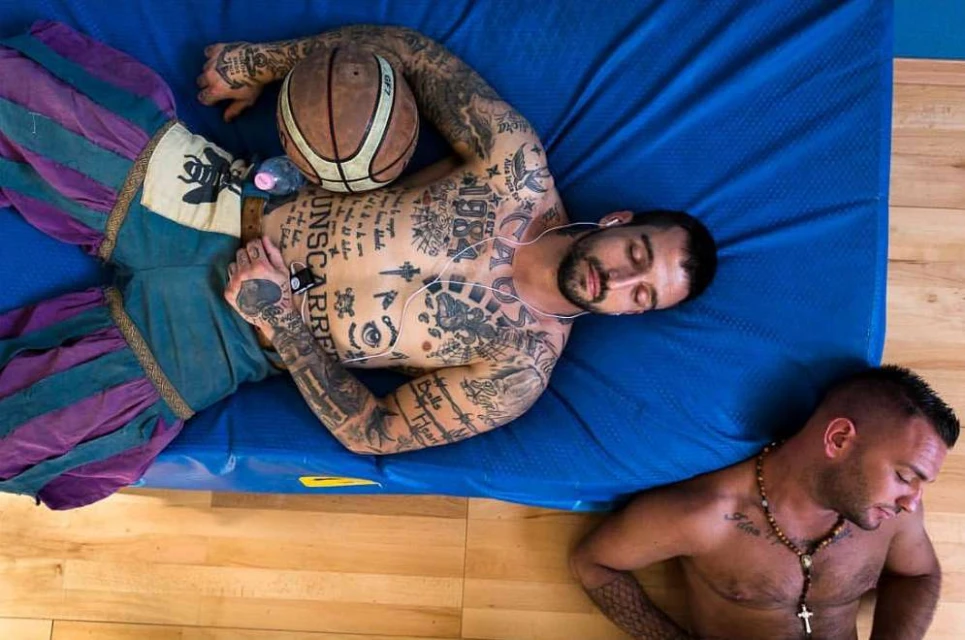Two shirtless men lie on a blue mat, one with tattoos and headphones next to a basketball, the other with a necklace and cross.