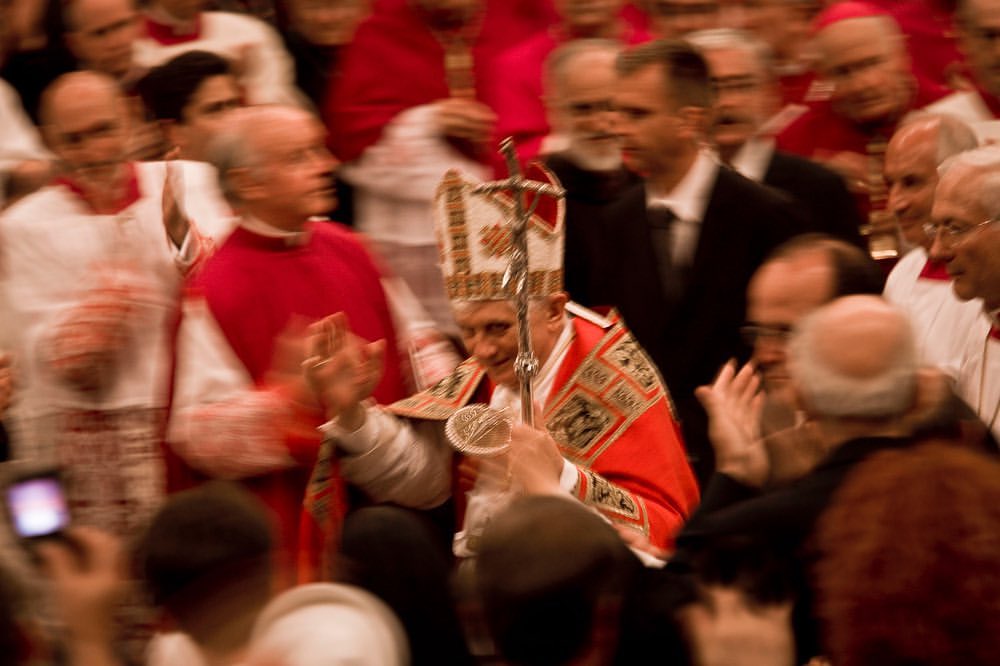 Crowd of people in an indoor setting, with a focus on a person wearing an ornate white and red robe and a matching hat.