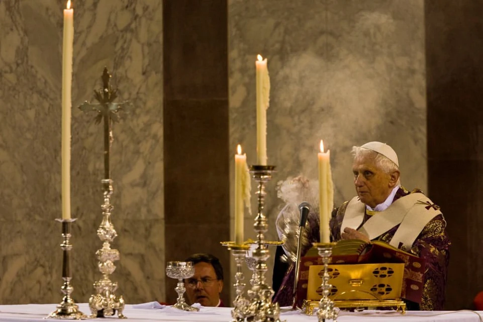 An adult male in religious attire officiating a ceremony with candles, a chalice, and a crucifix on a table.